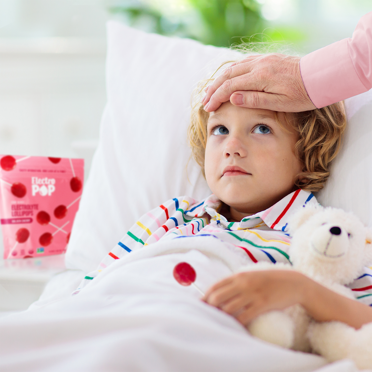 A child with curly hair, wearing striped pajamas, rests in bed under a white blanket, holding an electrolyte lollipop and a teddy bear. A hand gently touches the child's forehead, and a package of "ElectroPop" electrolyte lollipops is visible on the bedside table, suggesting care and hydration during illness.