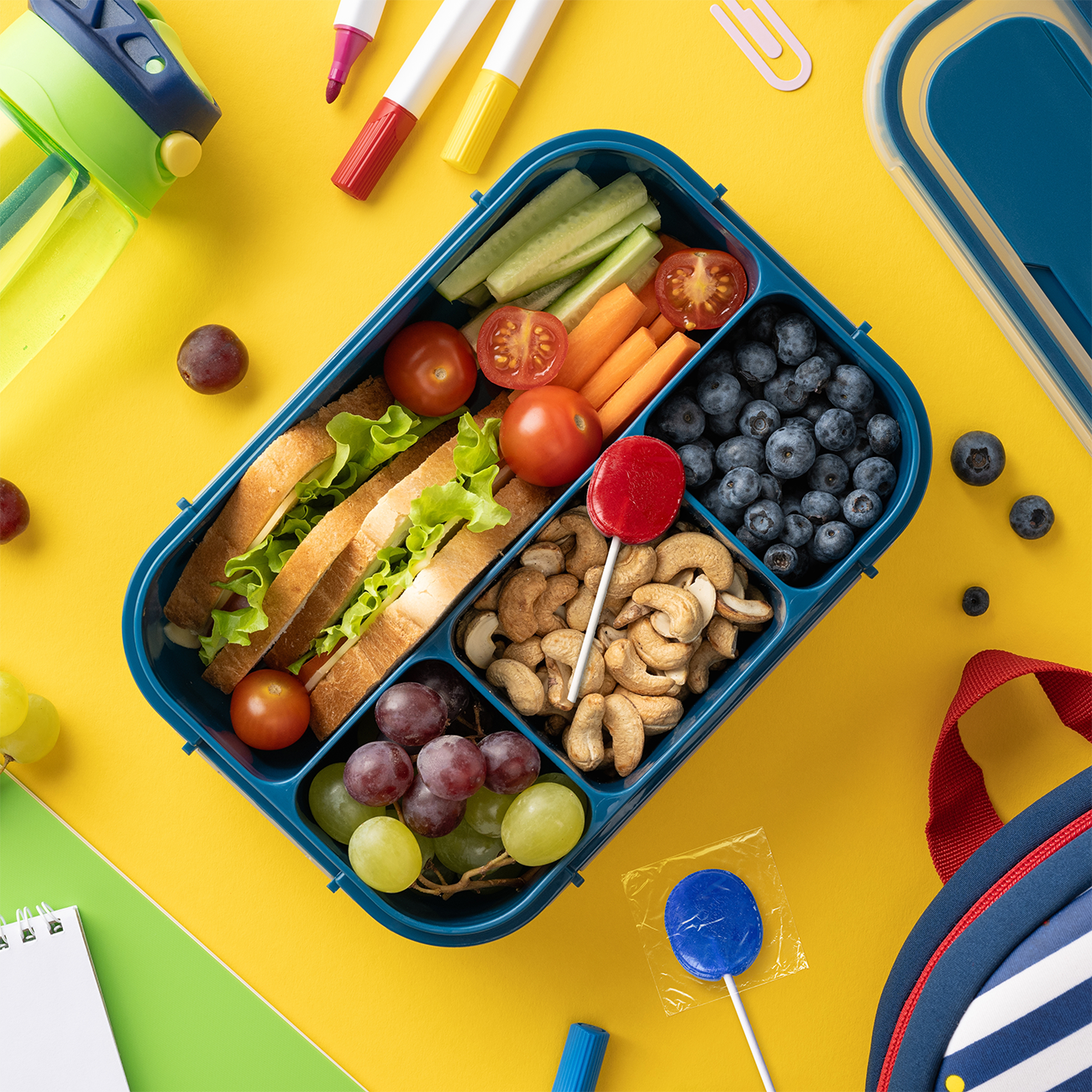 Healthy lunch box with electrolyte lollipop, sandwich, berries, and veggies on a yellow background with school supplies.